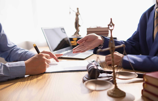 Two people in suits sit at a desk with legal documents, a laptop, a gavel, and a justice statue, suggesting a legal consultation or meeting in an office setting.