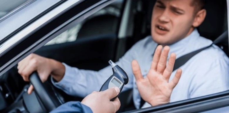 A man sitting in a car with one hand on the steering wheel raises his other hand to refuse a breathalyzer test being offered by someone outside the car.