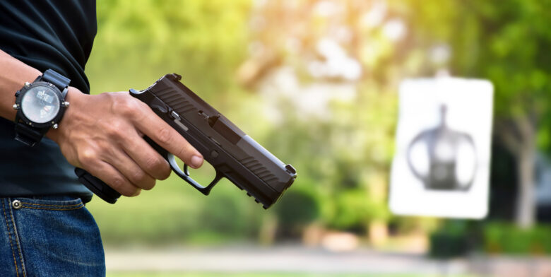 A person holding a handgun outdoors with a blurred target in the background. The person is wearing a black shirt, jeans, and a wristwatch. The scene suggests a shooting practice or firearm training session.